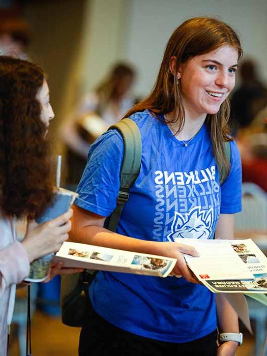 A smiling Billiken with long hair holds an admission booklet